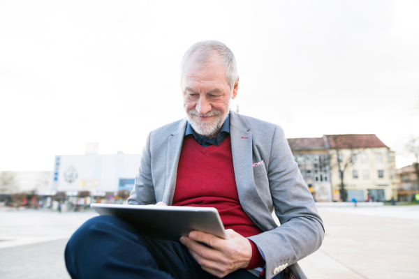 Handsome senior man in town sitting on bench, working on tablet.