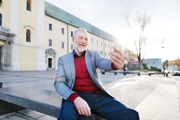 Handsome senior man in town sitting on bench, holding smart phone, taking selfie. Sunny spring day.