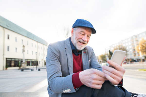 Handsome senior man in town sitting on bench, holding smart phone, texting. Sunny spring day.