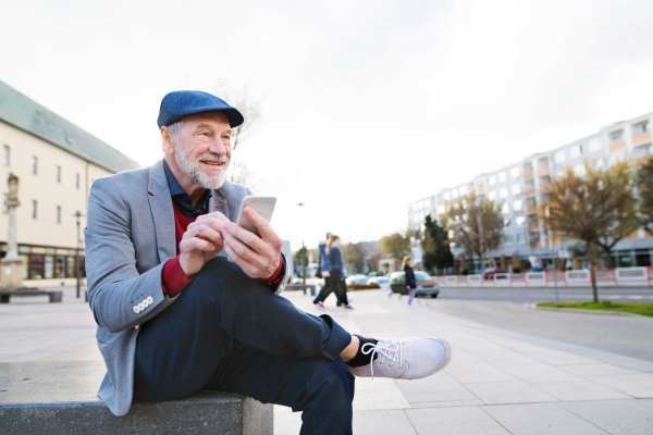 Handsome senior man in town sitting on bench, holding smart phone, texting. Sunny spring day.