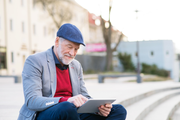 Handsome senior man in town sitting on stairs, holding laptop, smiling. Sunny spring day.