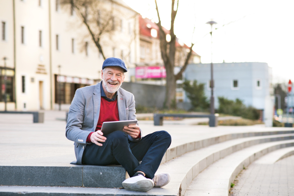 Handsome senior man in town sitting on stairs, holding laptop, smiling. Sunny spring day.