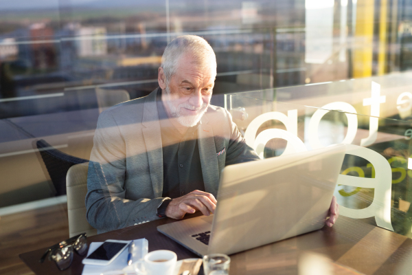 Handsome senior businessman working on laptop in cafe. Sunny spring day. View through glass.