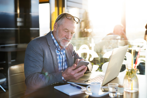 Handsome senior businessman with laptop in cafe, holding smart phone, texting. Sunny spring day.