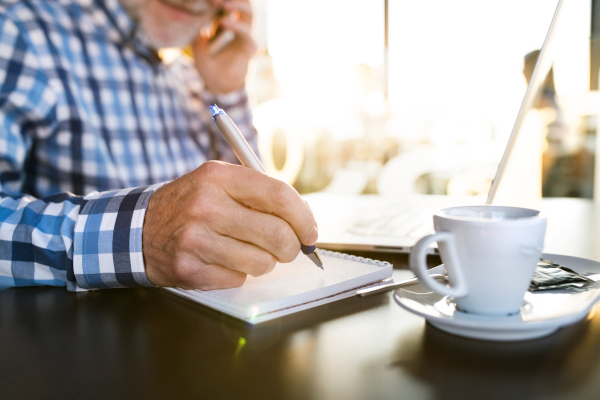 Unrecognizable senior businessman with laptop in cafe, holding smart phone, making phone call, writing something into his notebook. Sunny spring day.