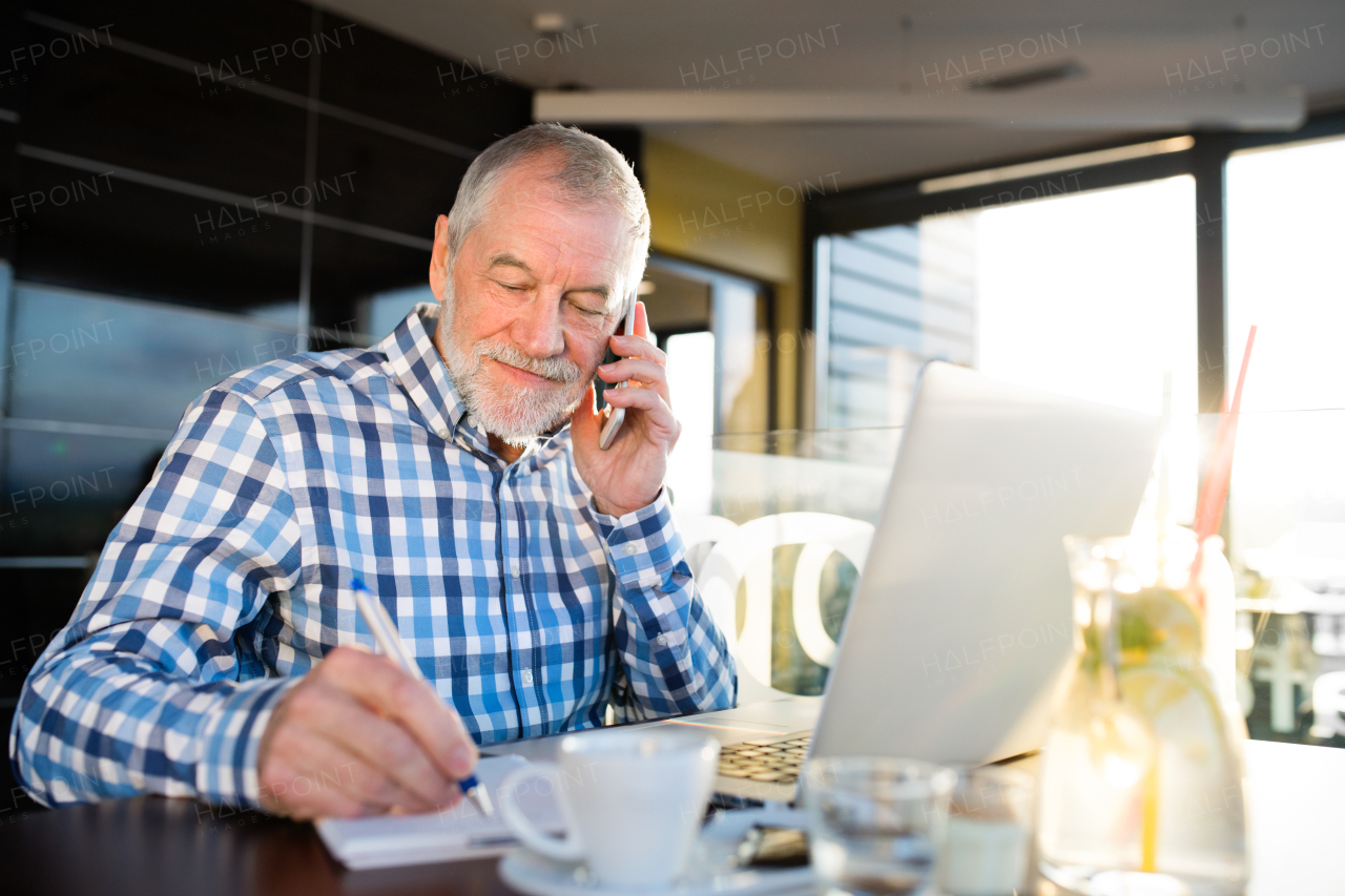 Handsome senior businessman with laptop in cafe, holding smart phone, making phone call. Sunny spring day.