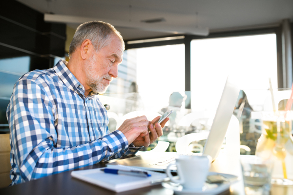 Handsome senior businessman with laptop in cafe, holding smart phone, texting. Sunny spring day.