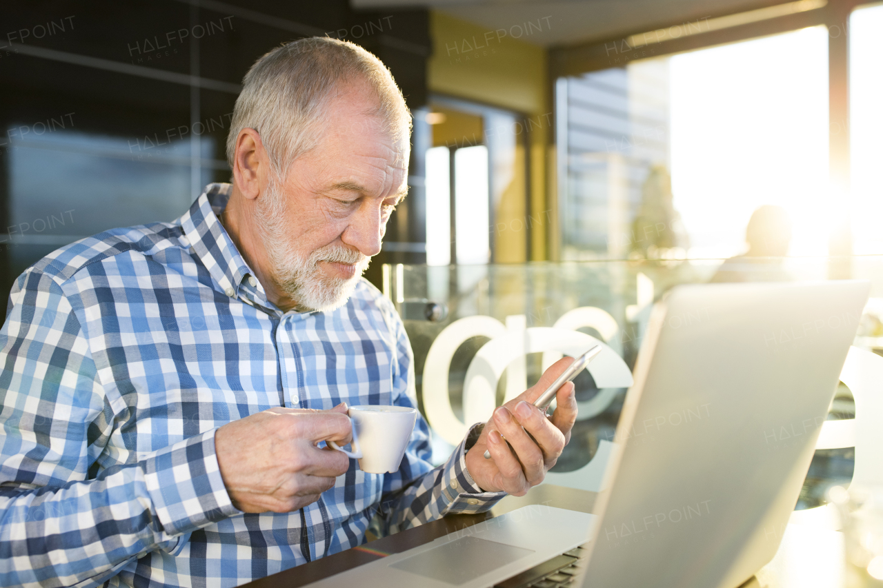 Handsome senior businessman with laptop in cafe, holding smart phone, reading or watching something. Sunny spring day.