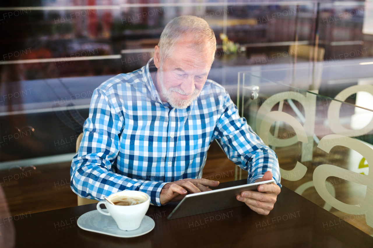 Handsome senior businessman working on tablet in cafe. Sunny spring day. View through glass.