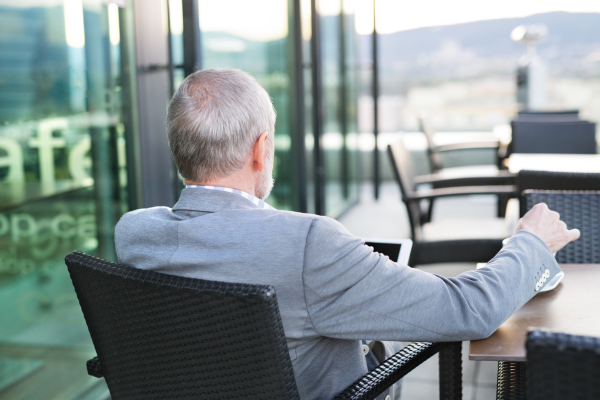 Handsome senior businessman with a tablet enjoying coffee in rooftop cafe. Sunny spring day. Rear view.