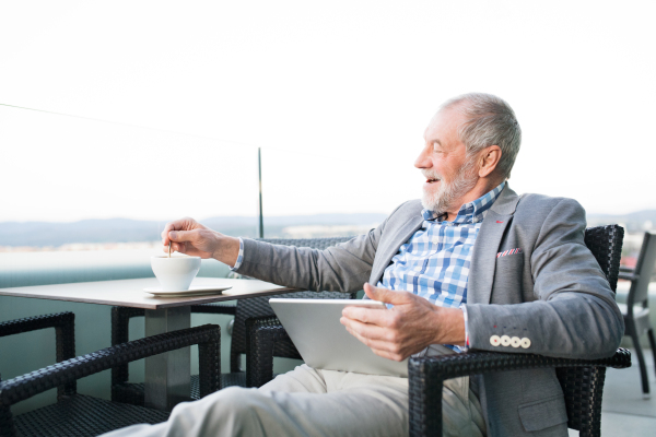 Handsome senior businessman working on tablet and enjoying coffee with his feet up in rooftop cafe. Sunny spring day.