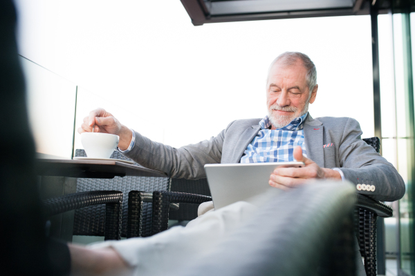 Handsome senior businessman working on tablet and enjoying coffee with his feet up in rooftop cafe. Sunny spring day.