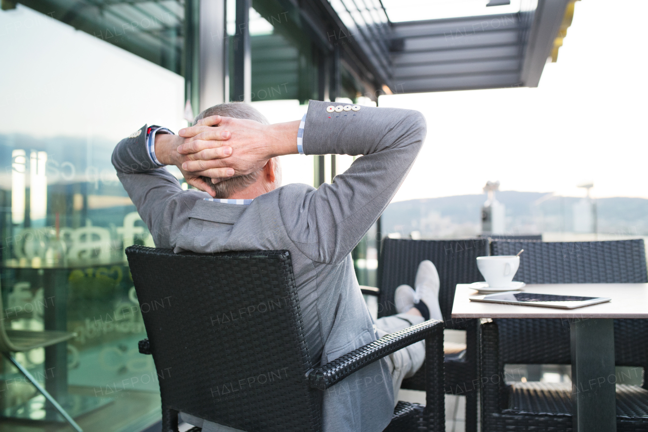 Handsome senior businessman with a tablet enjoying coffee in rooftop cafe. Sunny spring day. Rear view.