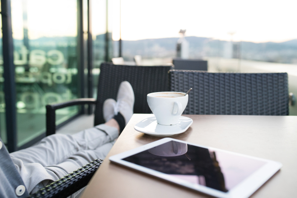 Unrecognizable businessman with a tablet enjoying coffee in rooftop cafe. Sunny spring day. Rear view.