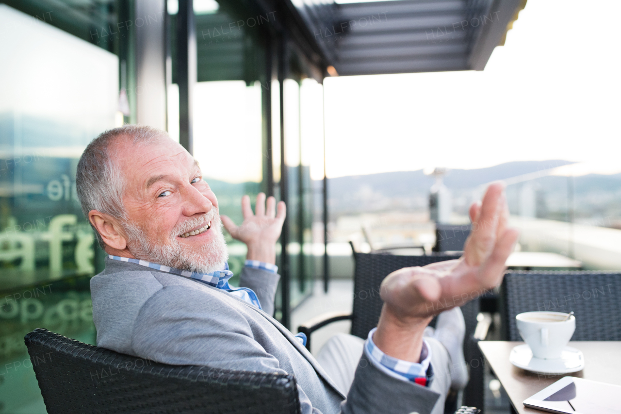 Handsome senior businessman with a tablet enjoying coffee in rooftop cafe. Sunny spring day. Rear view.