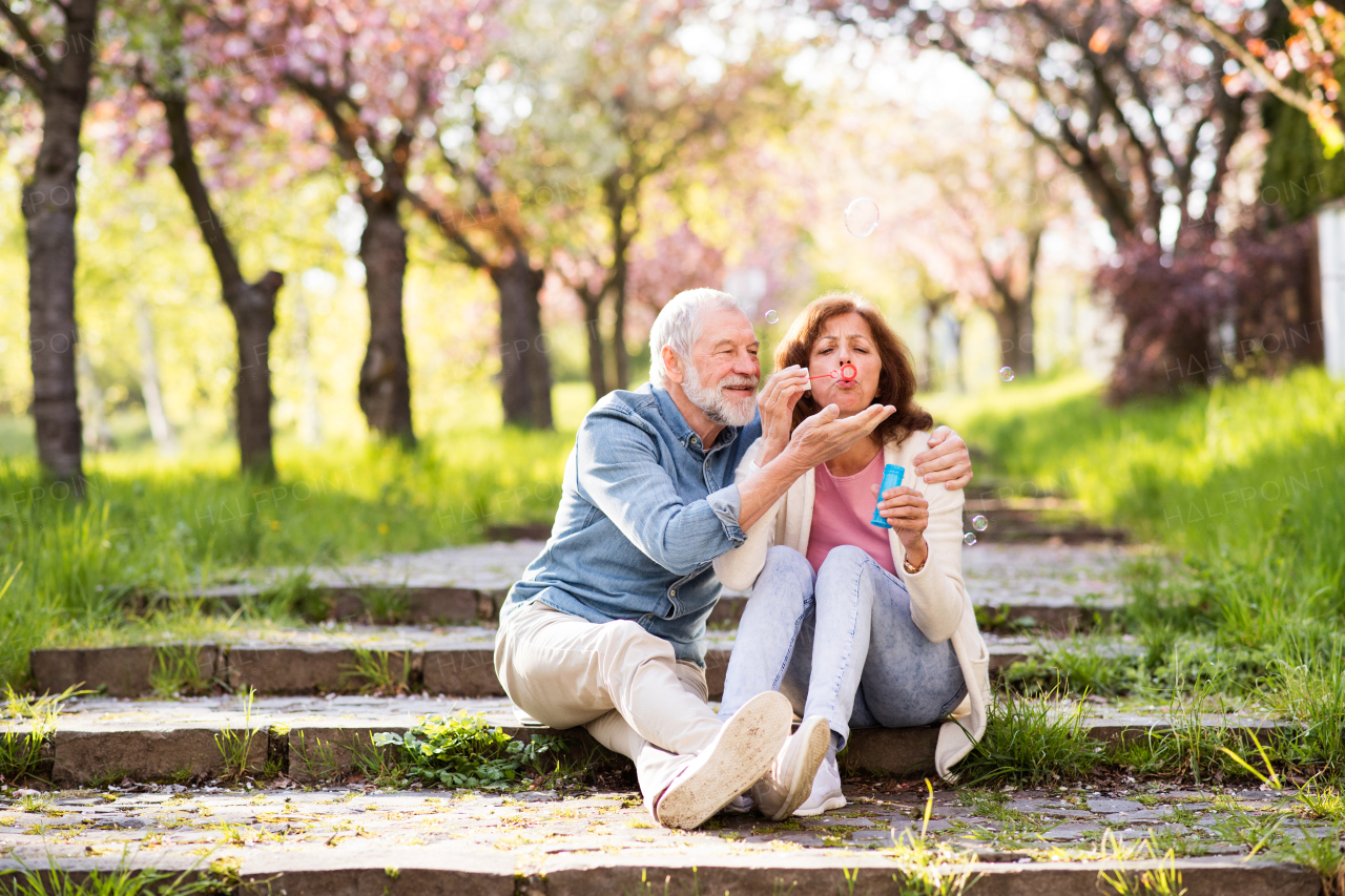 Beautiful senior couple in love outside in spring nature, sitting on stairs, blowing bubbles with bubble wand.