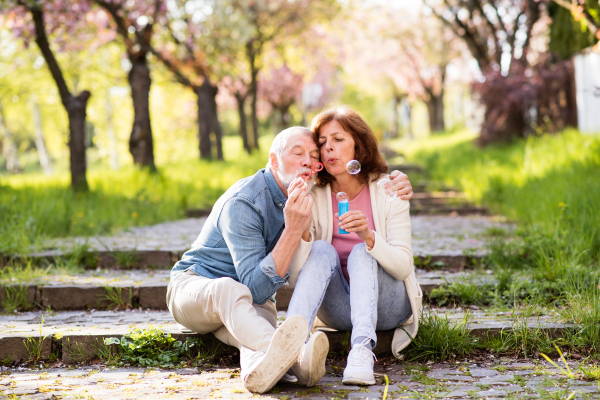 Beautiful senior couple in love outside in spring nature, sitting on stairs, blowing bubbles with bubble wand.