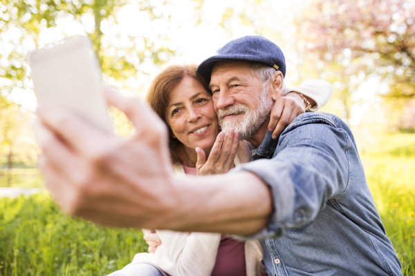 Beautiful senior couple in love on a walk outside in spring nature under blossoming trees. Man and woman taking a selfie with a smartphone.