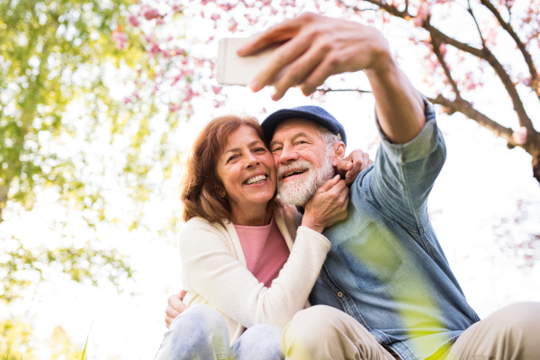 Beautiful senior couple in love on a walk outside in spring nature under blossoming trees. Man and woman taking a selfie with a smartphone.