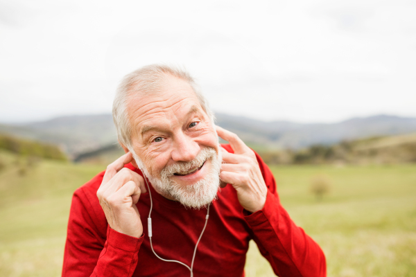 Active senior runner in red sweatshirt with earphones listening music outside on green hills, taking break, resting.