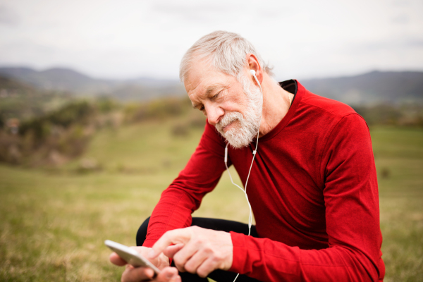 Active senior runner outside on green hills with smart phone and earphones, resting, listening music.