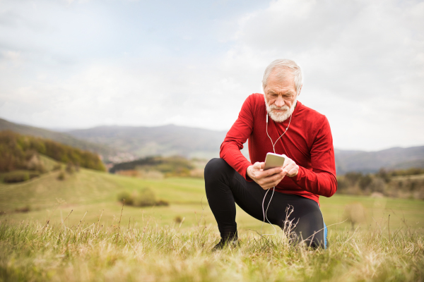 Active senior runner outside on green hills with smart phone and earphones, resting, listening music.