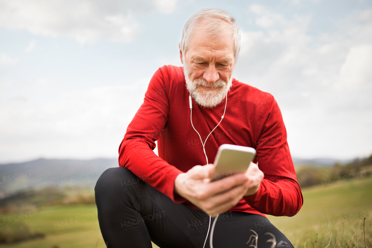 Active senior runner outside on green hills with smart phone and earphones, resting, listening music.