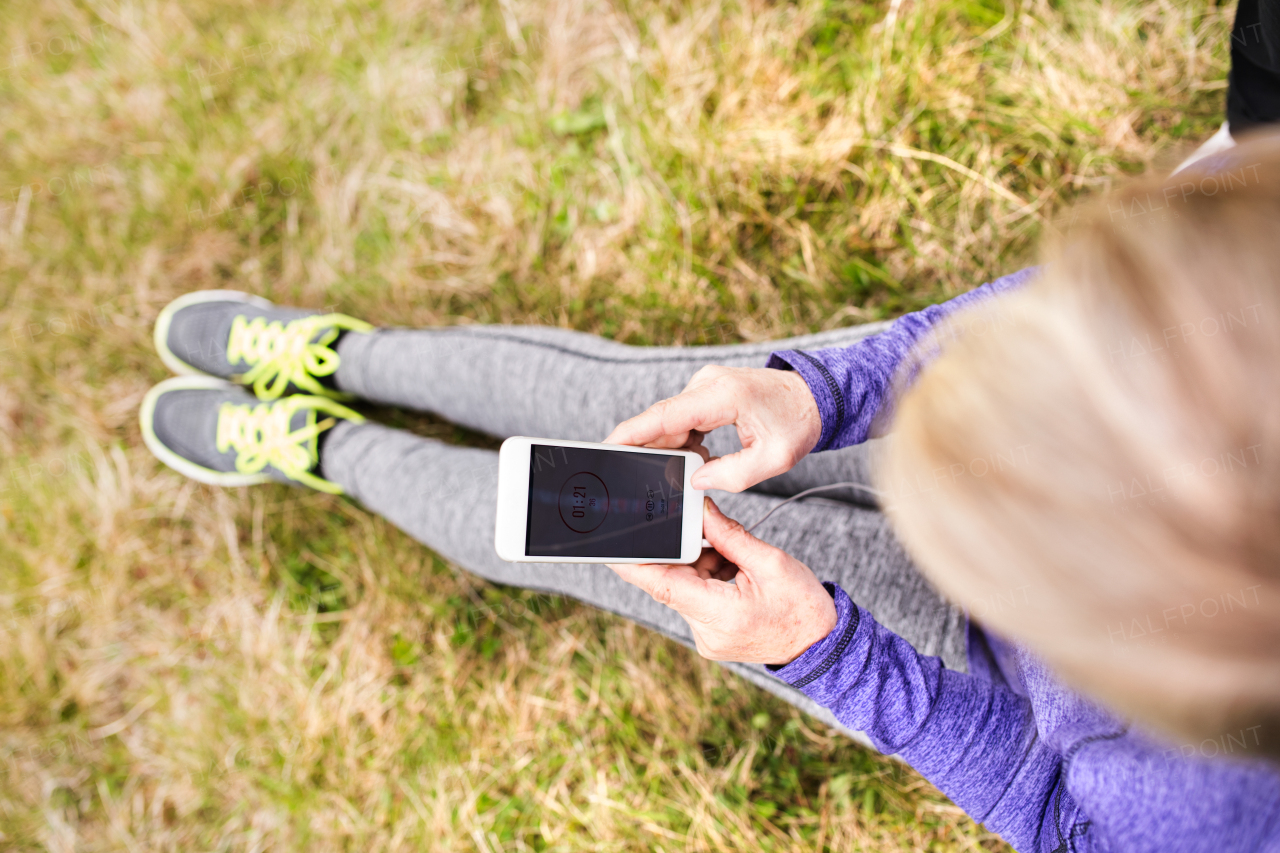 Unrecognizable active senior runner with smart phone outside in nature sitting on the grass.