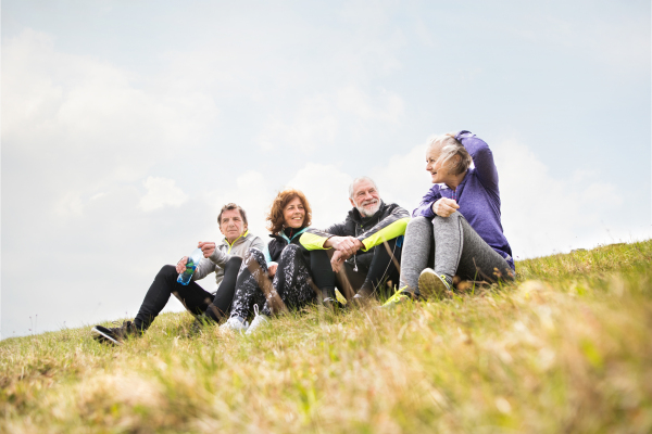 Group of active senior runners outdoors, sitting on the grass, resting and talking together. Rear view.