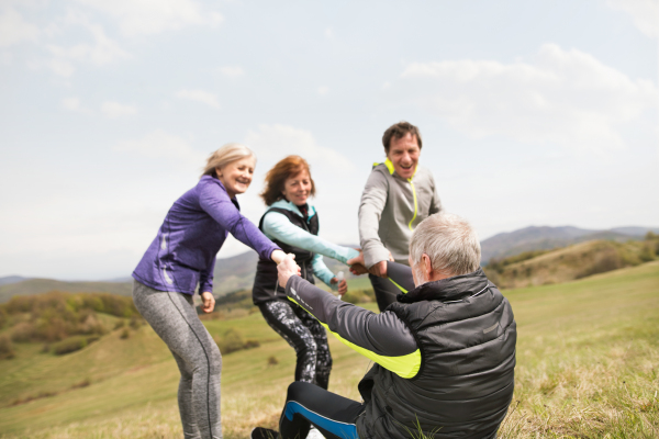 Group of active senior runners outdoors, sitting on the grass, resting and talking together, helping senior man to stand up.