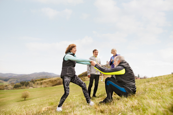 Group of active senior runners outdoors, sitting on the grass, resting and talking together, helping senior man to stand up.