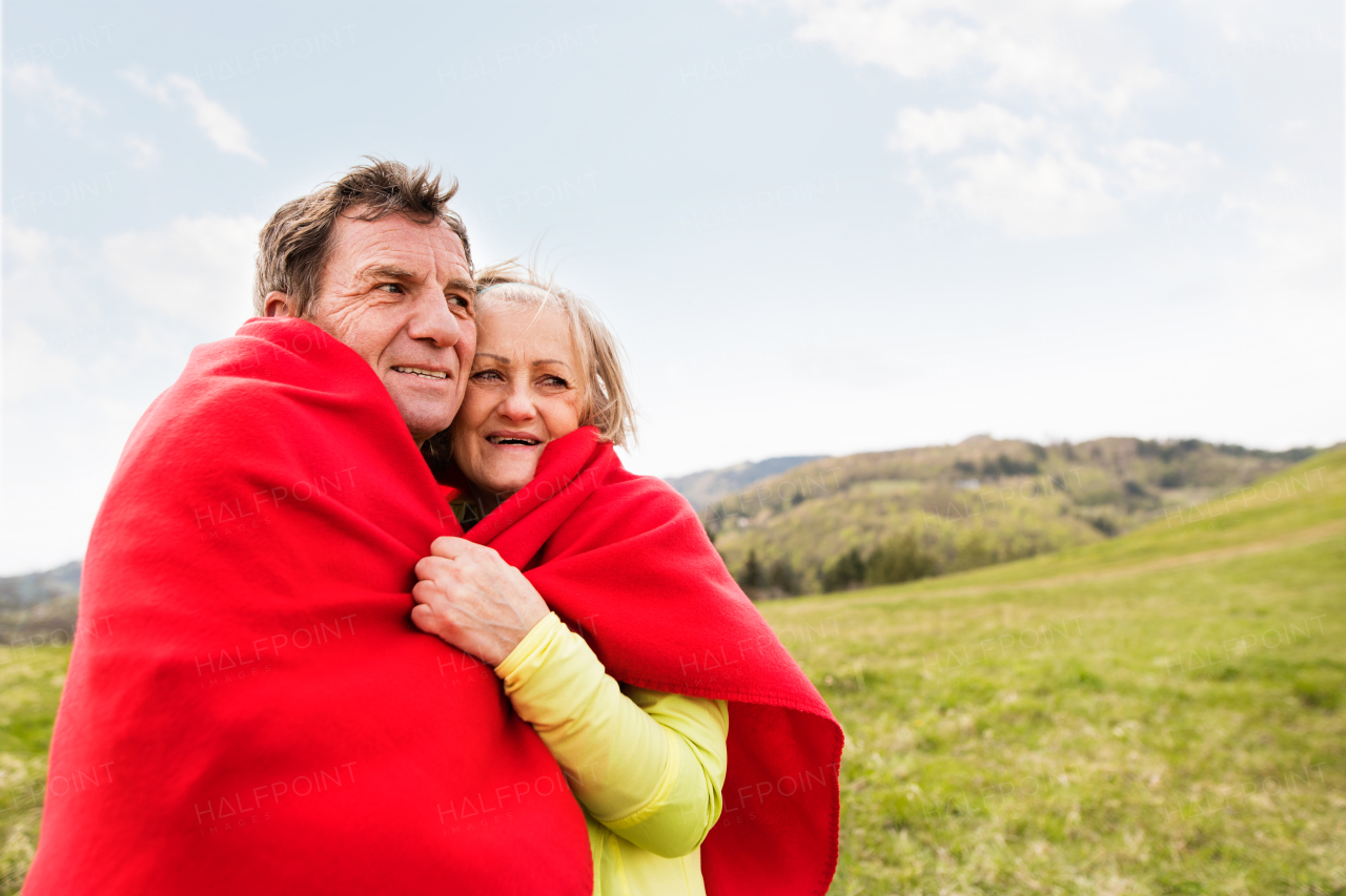 Beautiful active senior runners resting outside in sunny autumn nature, wrapped in red blanket, hugging.