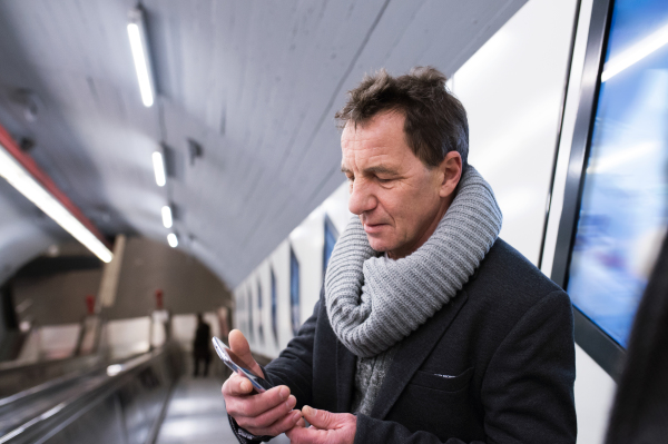 Senior man in winter clothes holding a smart phone, standing at the escalator in Vienna subway