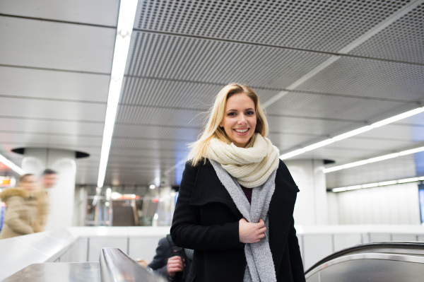 Beautiful young blonde woman in black coat and big woolen scarf standing at the escalator in Vienna subway, holding shopping bags