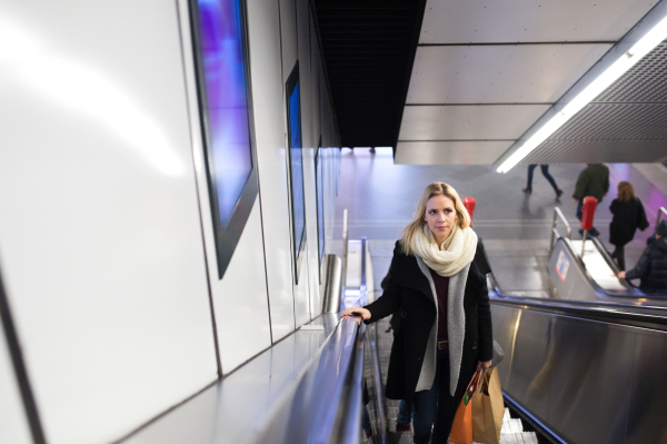 Beautiful young blonde woman in black coat and big woolen scarf standing at the escalator in Vienna subway, holding shopping bags