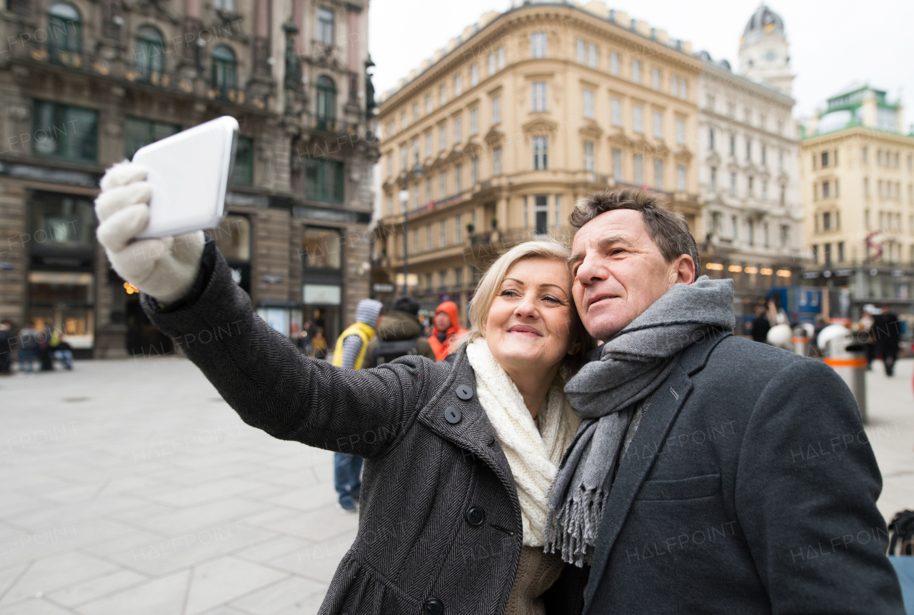 Beautiful senior couple in winter clothes on a walk in historical centre of the city of Vienna, Austria. Woman taking selfie of them with smart phone.