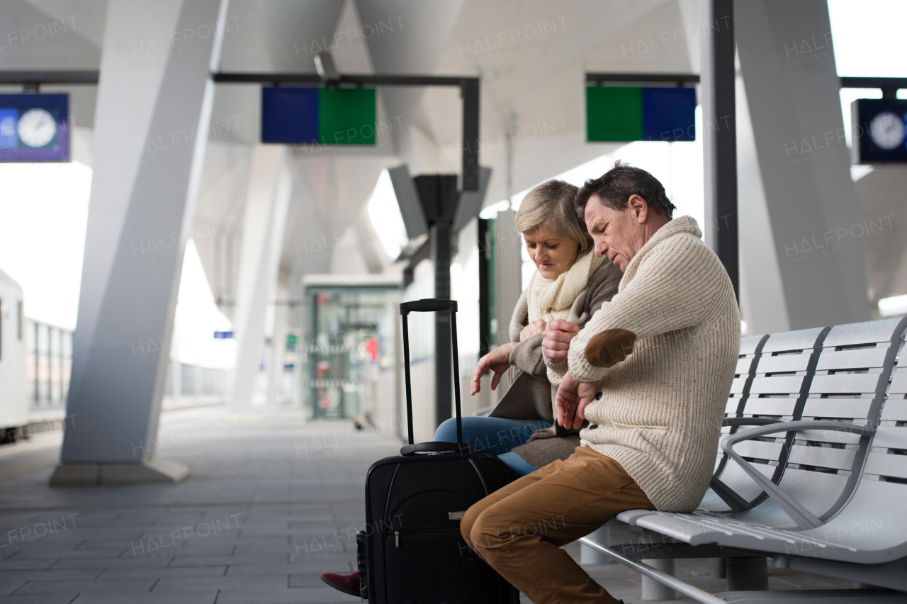 Beautiful senior couple waiting on train station, sitting on bench, looking at watch.