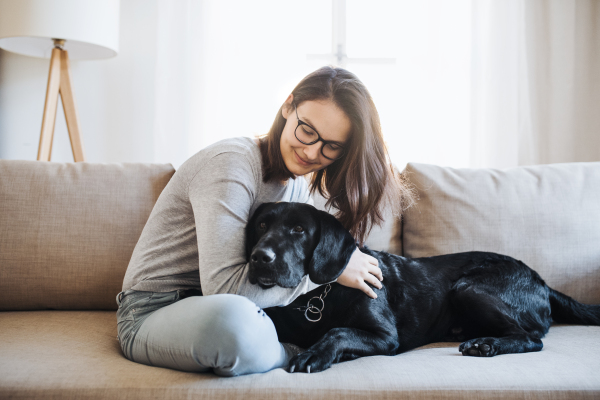 Teenage girl sitting on a sofa indoors, playing with a black pet dog.