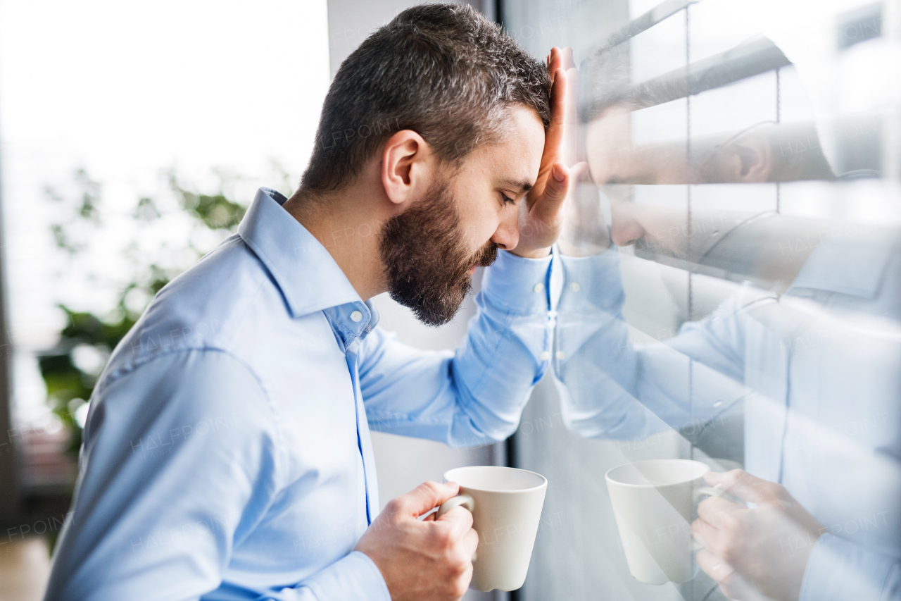 An unhappy man standing by the window, holding a cup of coffee.