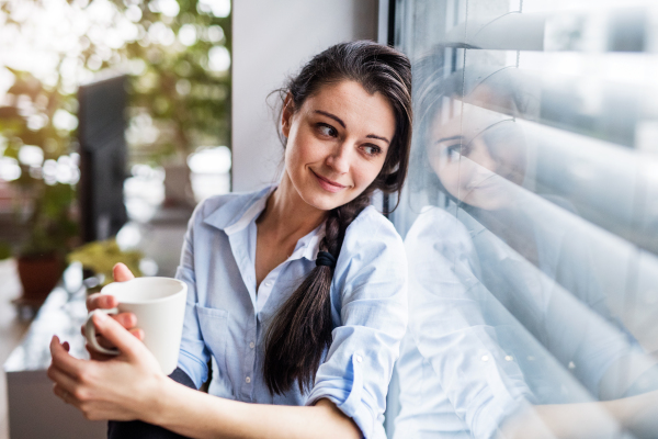 A woman sitting by the window, holding a cup of coffee. Smart home control system.