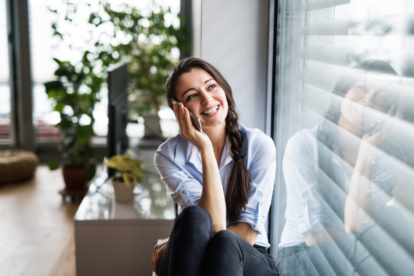 An attractive woman with smartphone sitting by the window at home, making a phone call.