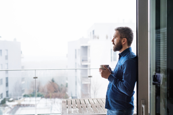 A man standing by the window, holding a cup of coffee at home.