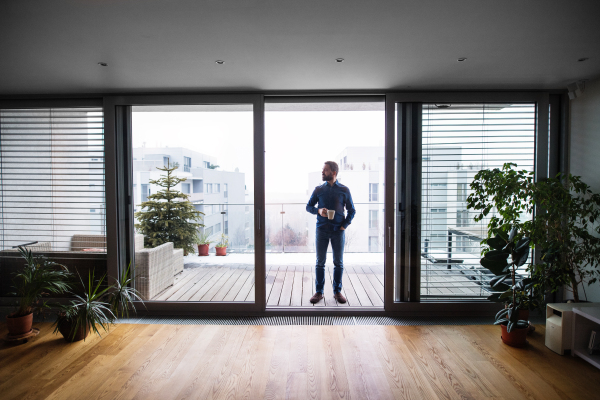 A man standing by the window on a balcony, holding a cup of coffee at home.