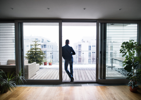 A man standing by the window, holding a cup of coffee at home. Rear view.