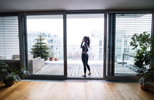 A woman standing by the window, holding smartphone and making a phone call.