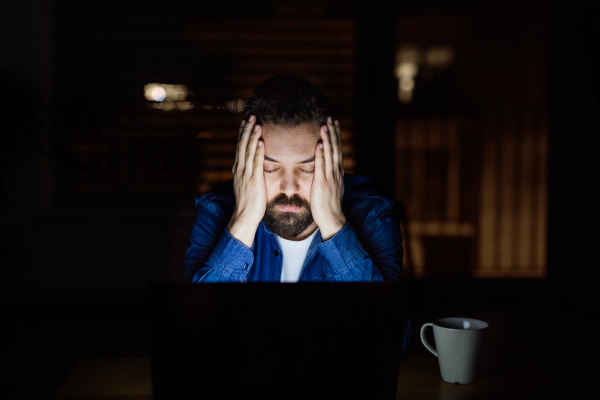 A tired man working on a laptop at home at night.