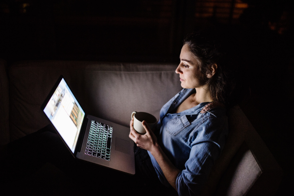 A woman sitting on a sofa at night, working on a laptop.