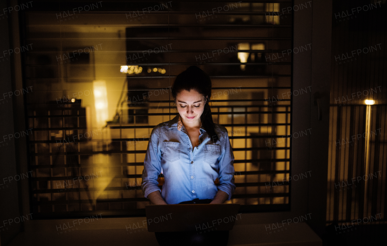 A woman sitting on a window sill at night, working on a laptop at home or in the office.