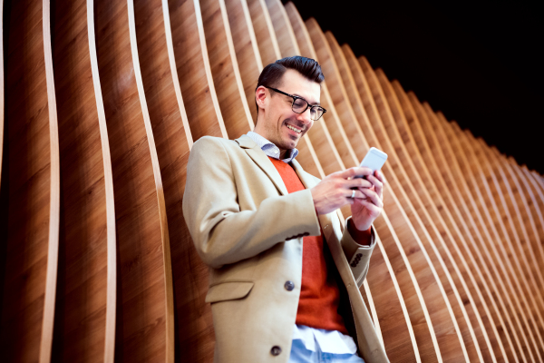 Young handsome man with smartphone standing against wooden wall, texting.