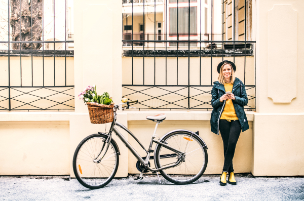 Portrait of a young beautiful woman with bicycle and smartphone standing against a building. Sunny spring town.